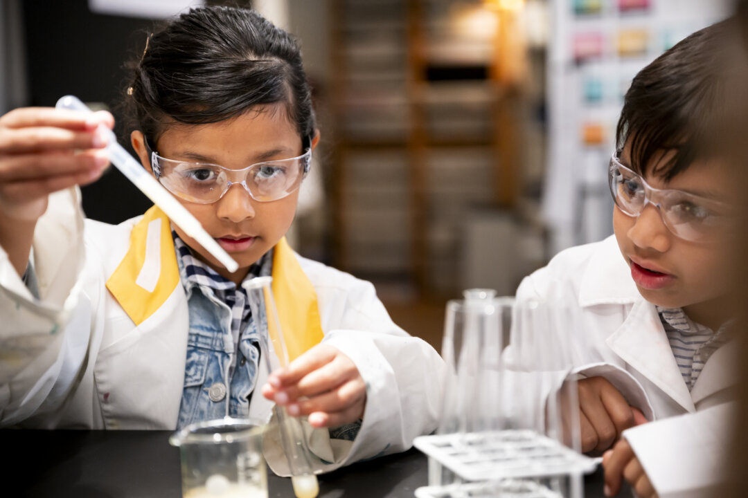 Two children in lab coats and safety goggles are conducting a chemistry experiment using a pipette.